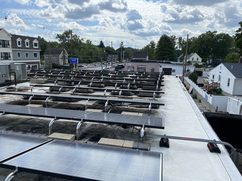 Rooftop view of a solar panel installation on a flat commercial roof: rows of panels on tilted racking with mounting rails and ballast blocks, some panels partially installed with wiring exposed, neighborhood houses, a 'Sunnyside Liquors' sign, and trees in the background.