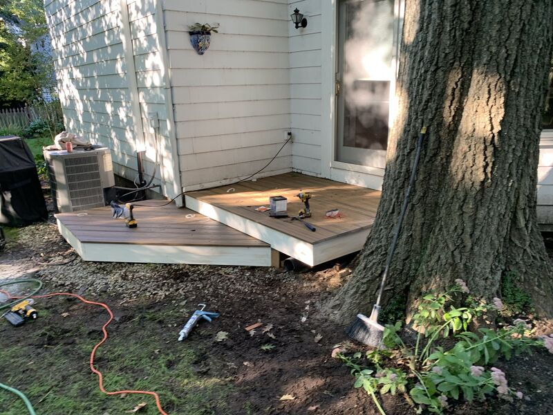 New wood-composite deck under construction against the back of a house, deck boards laid, cordless drill and driver bit box sitting on the deck, AC condenser unit and a large tree trunk beside the work area, garden hose in the yard.