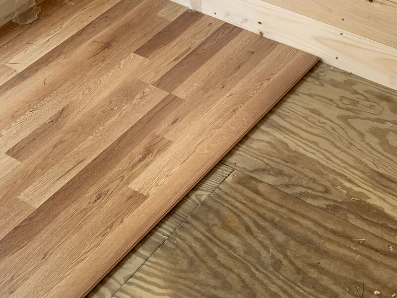 Close-up of new light hickory-tone laminate/engineered wood flooring being installed over a plywood subfloor, one finished row of planks laid next to exposed subfloor and fresh baseboard trim.