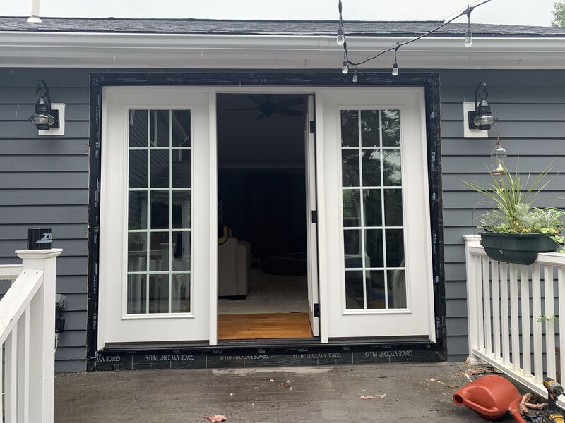 Finished double French doors with 15-lite glass panels opening onto a covered deck, black lantern sconces on either side, string lights overhead, dark blue-gray siding, hanging planter, and a bright interior room with wood flooring visible through the doors.