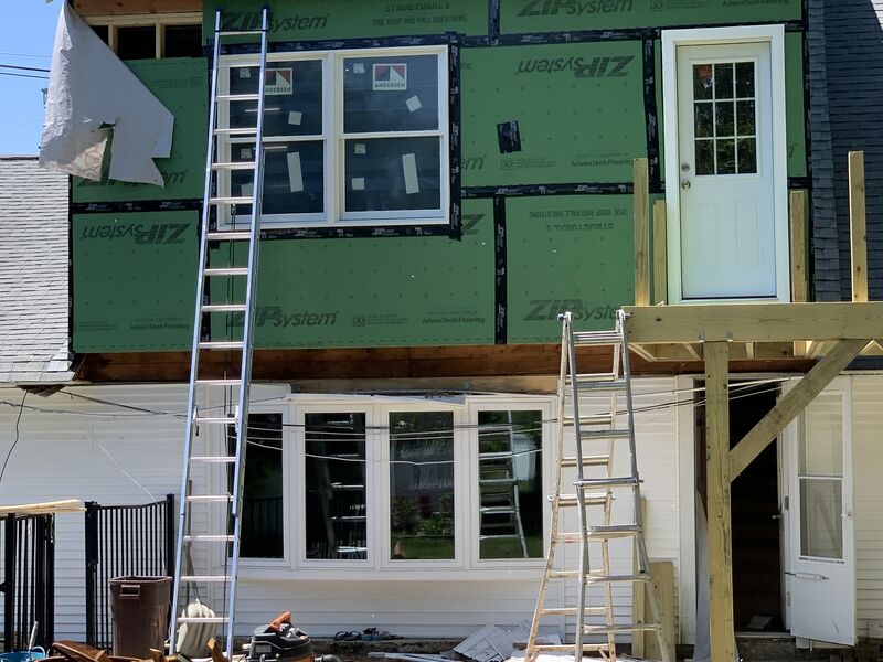 Two-story house addition mid-construction: green ZIP System structural sheathing panels with taped seams, two new Andersen windows and a white exterior door installed above, two extension ladders leaning against the wall, older vinyl-sided section of the house below with white bay windows.