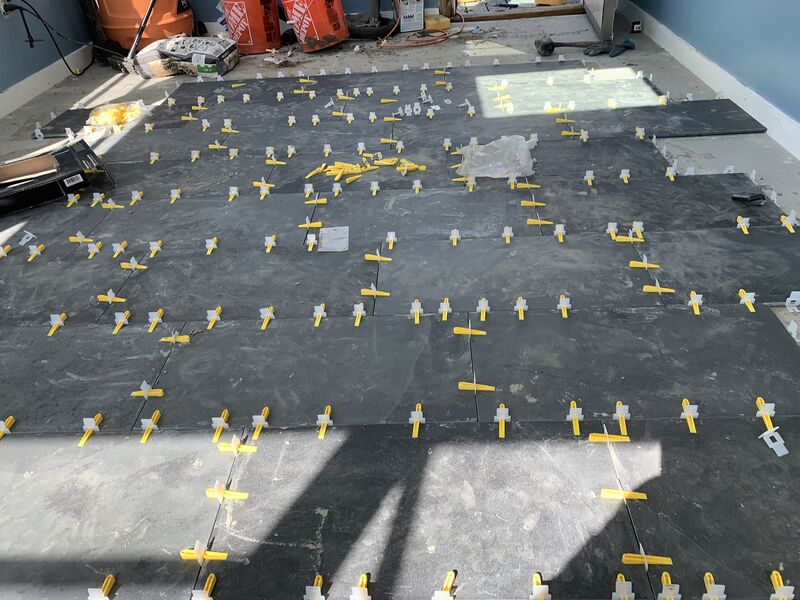 Interior room mid-tile-installation: dark gray large-format floor tiles laid with yellow tile-leveling spacer clips across the surface, Home Depot buckets and tile boxes stacked in the corner, blue-painted walls with white baseboard.