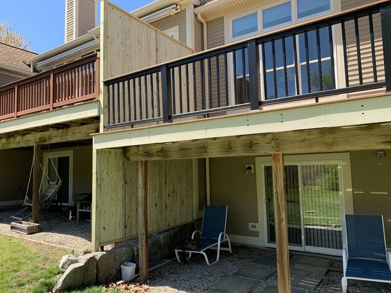Two-story house exterior with an upper wood-frame deck under construction, black metal railing balusters, and a natural wood privacy panel; a covered patio below has a porch swing, patio chairs, and sliding glass doors.